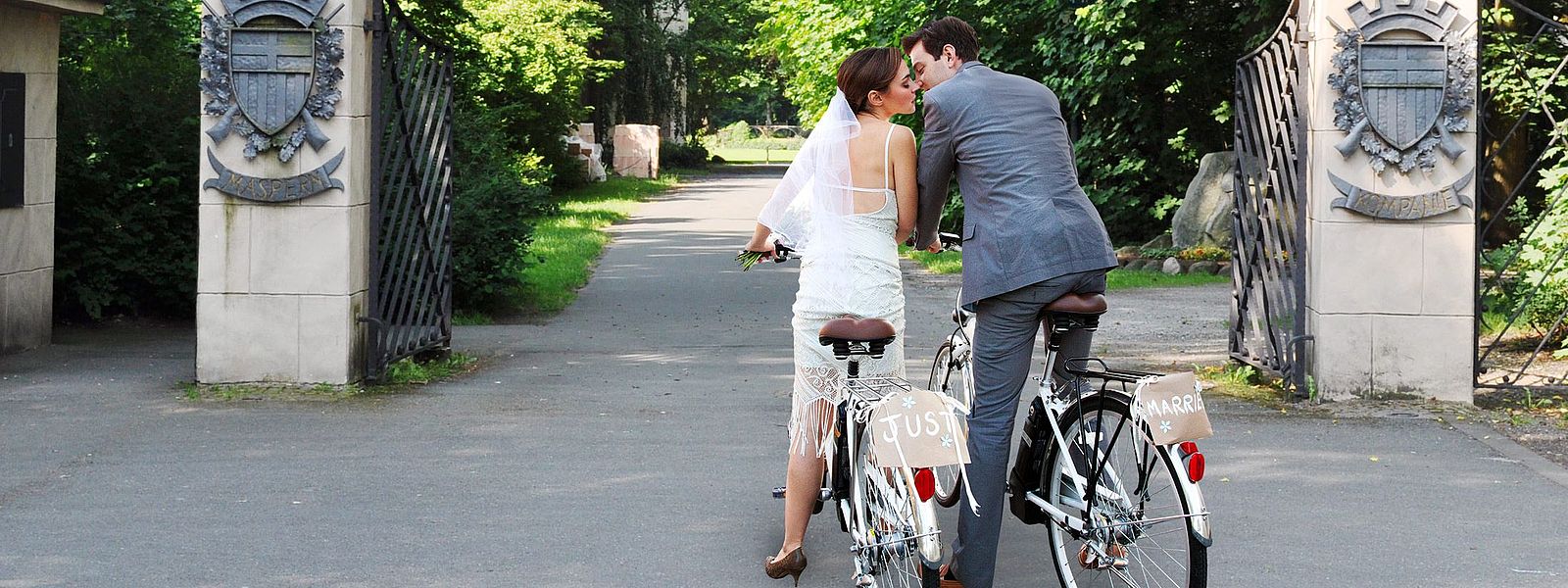 Hochzeitspaar in Brautkleid und Anzug auf zwei Fahrrädern vor den Toren zum Schützenplatz
