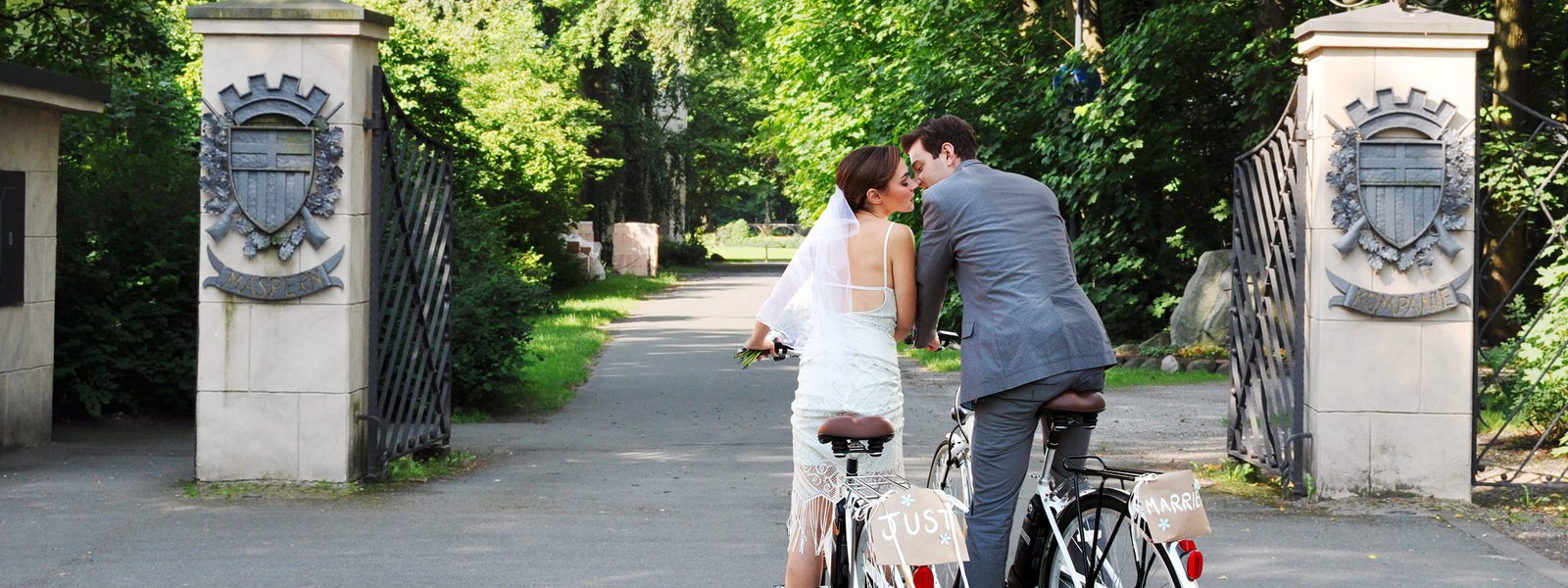 Hochzeitspaar in Brautkleid und Anzug auf zwei Fahrrädern vor den Toren zum Schützenplatz