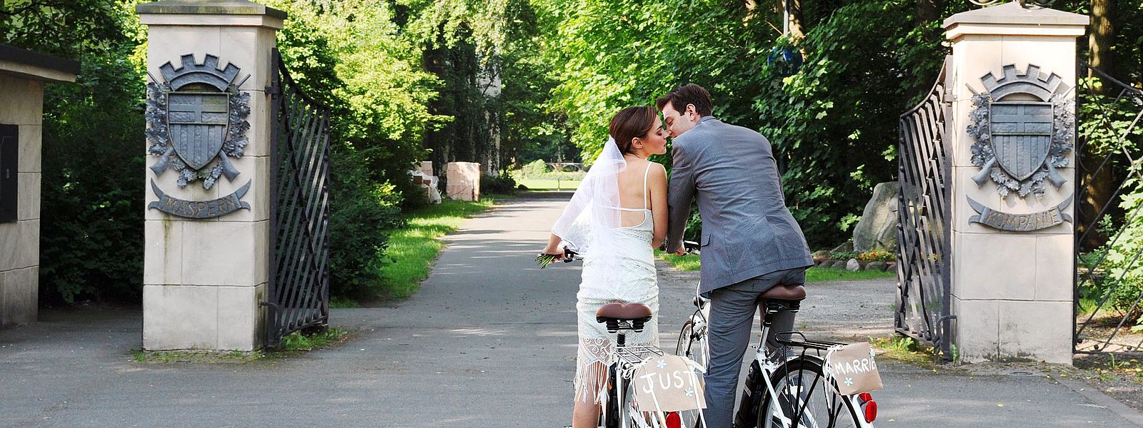 Hochzeitspaar in Brautkleid und Anzug auf zwei Fahrrädern vor den Toren zum Schützenplatz