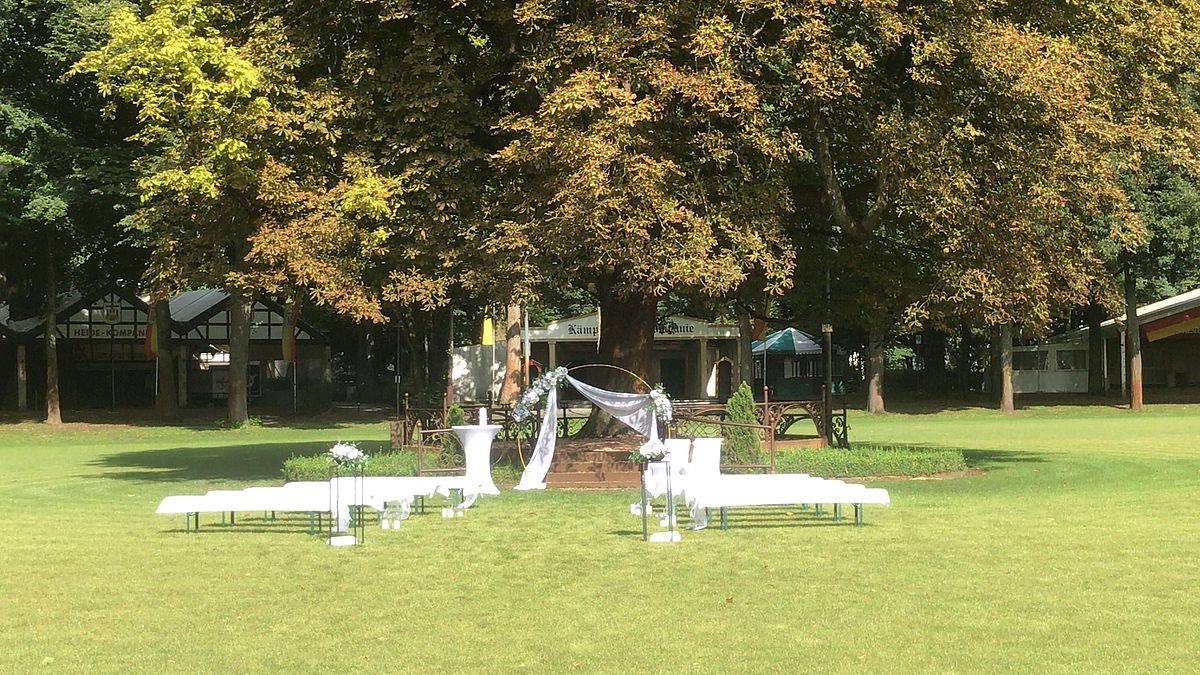 Foto der Kastanie in der Mitte des grünen Schützenplatzes mit weißen Bänken und Hochzeitsbogen darunter