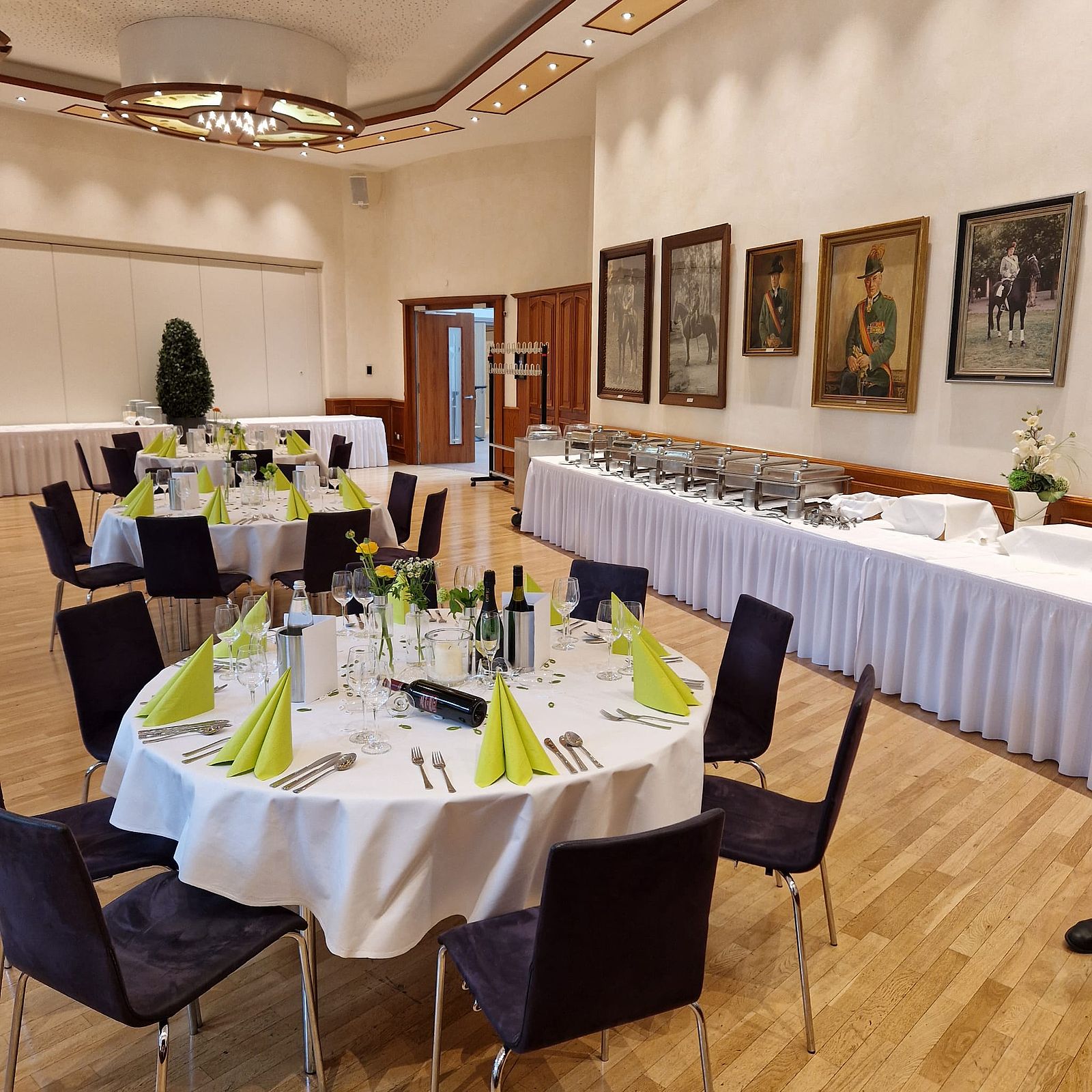Ausschnitt des Fahnensaals mit runden Tischen und grüner Deko, im Hintergrund der Aufbau fürs Buffet