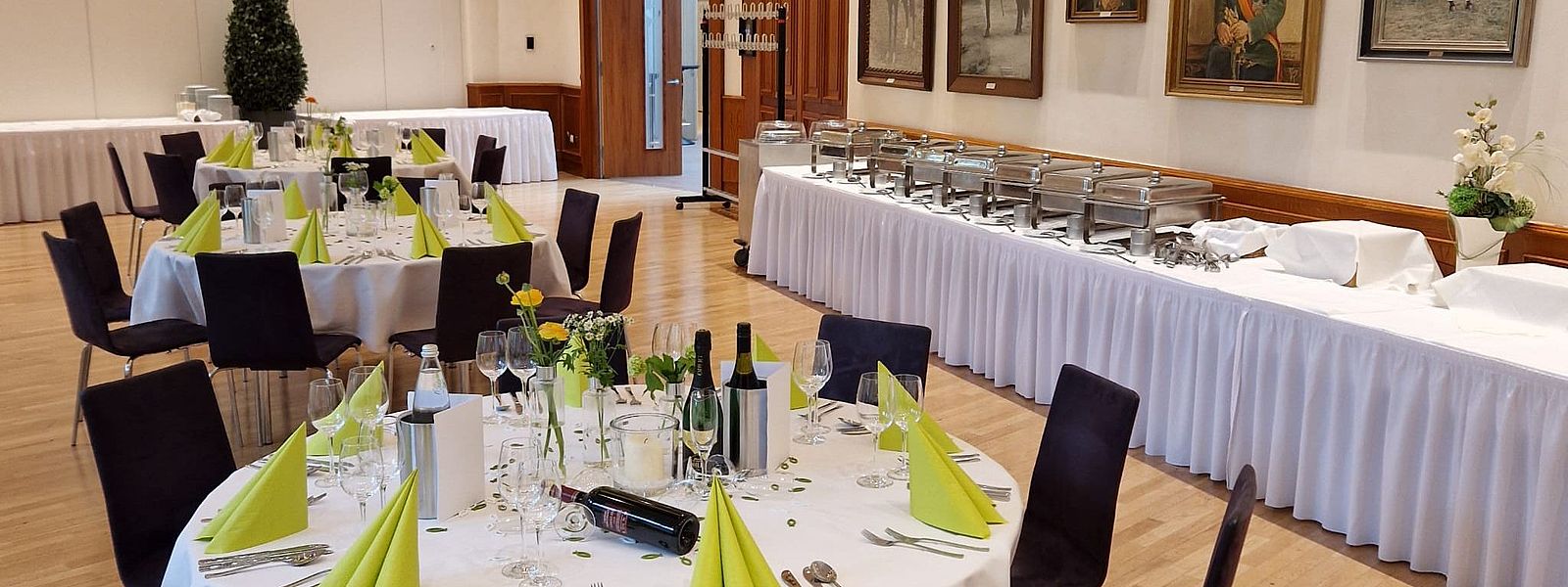 Ausschnitt des Fahnensaals mit runden Tischen und grüner Deko, im Hintergrund der Aufbau fürs Buffet
